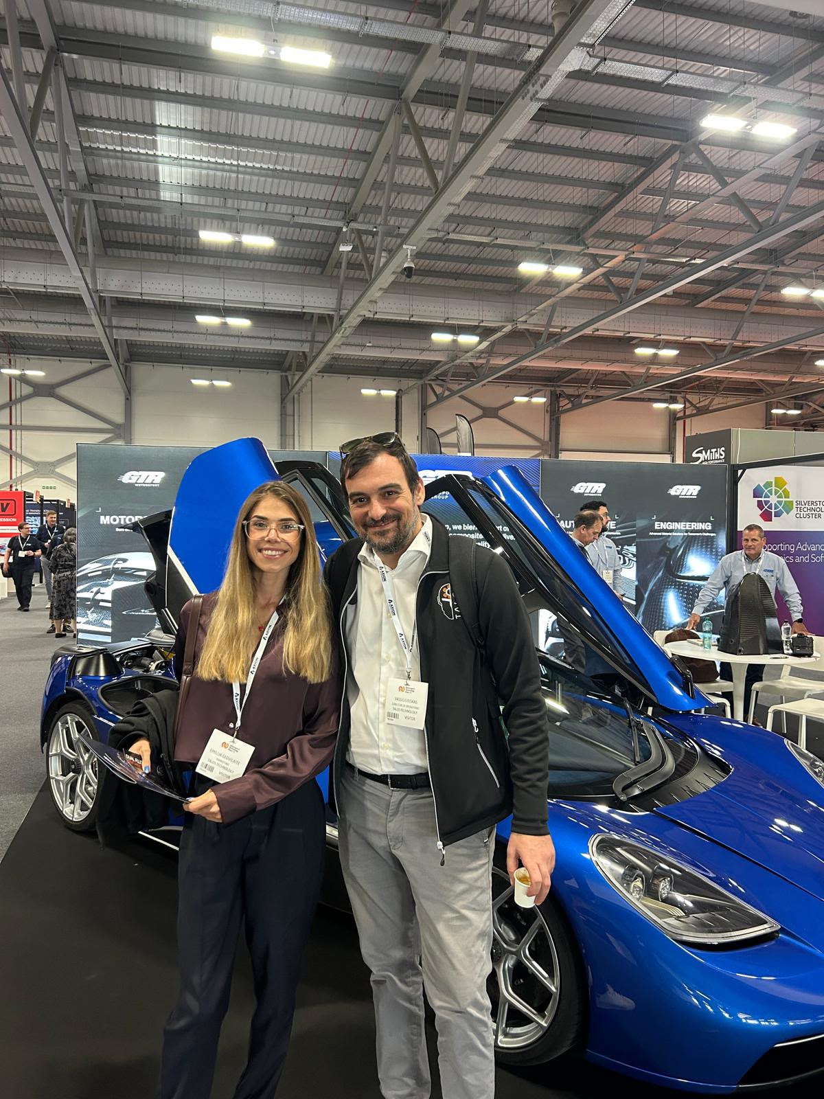 Emilija Gedvilaite and Vassilis Riskas of Talos Technology beside a blue sports car at Motorsport Technology Expo UK 2026